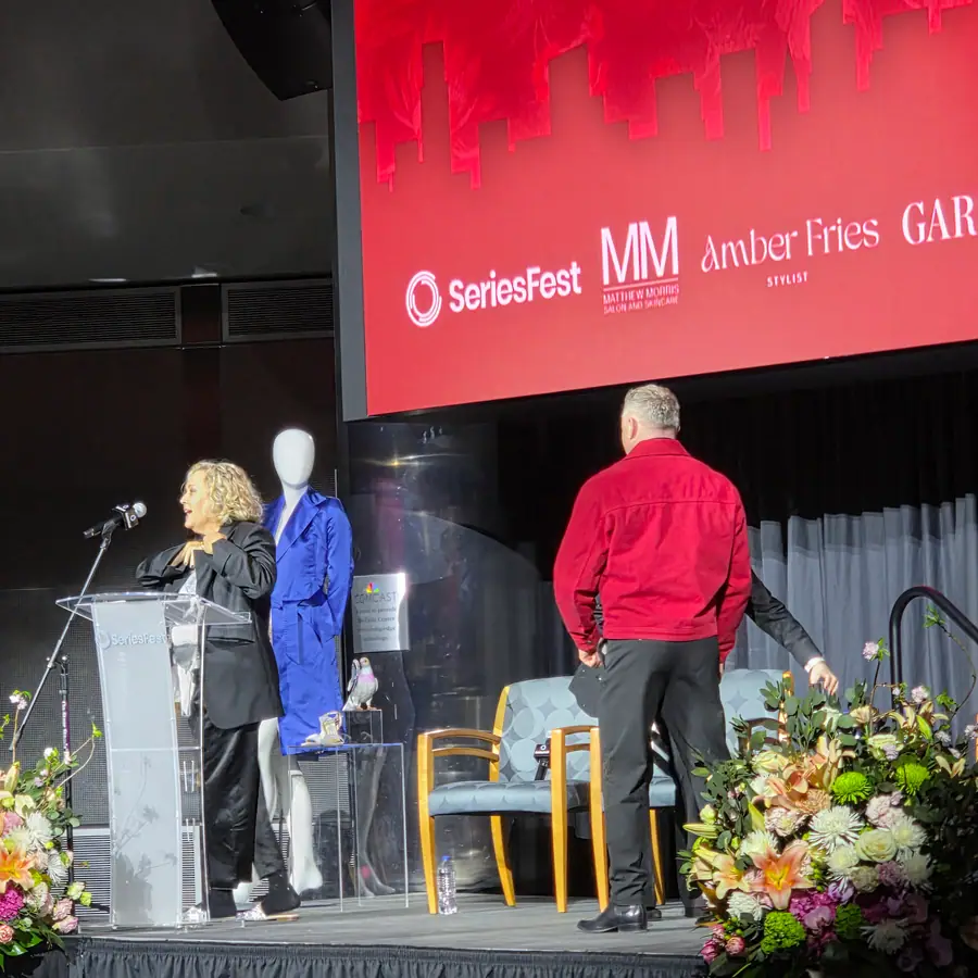 A host at the podium beside the blue-coat mannequin, the MM · Amber Fries · Garbarini sponsor screen behind.