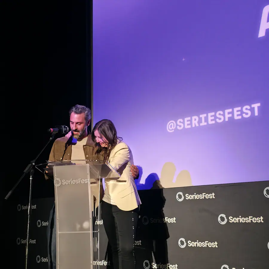 Two people at the SeriesFest podium, mid-embrace, the branded screen behind them.