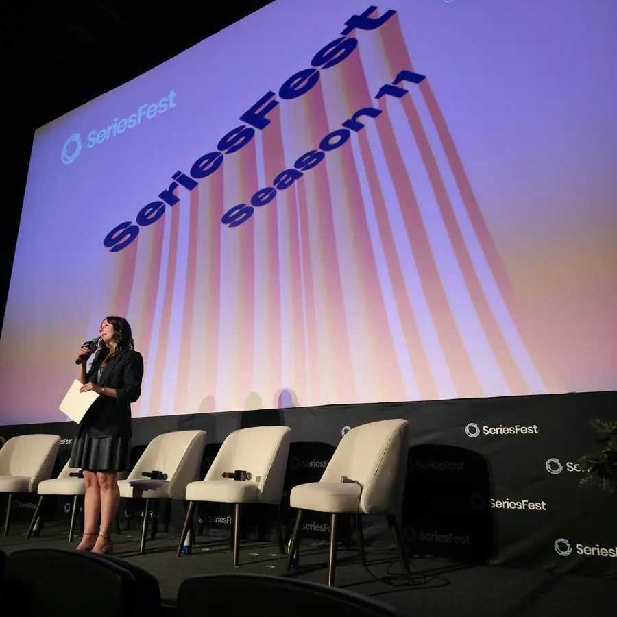 A moderator speaks from the SeriesFest podium, notecard in hand, before an empty panel row and the Season 11 branded screen.