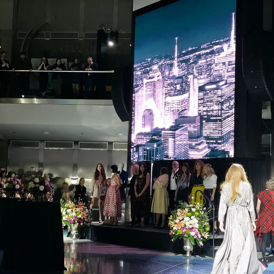 A model in a plaid coat walks the runway past a floral arrangement, a big screen behind displaying a night skyline.