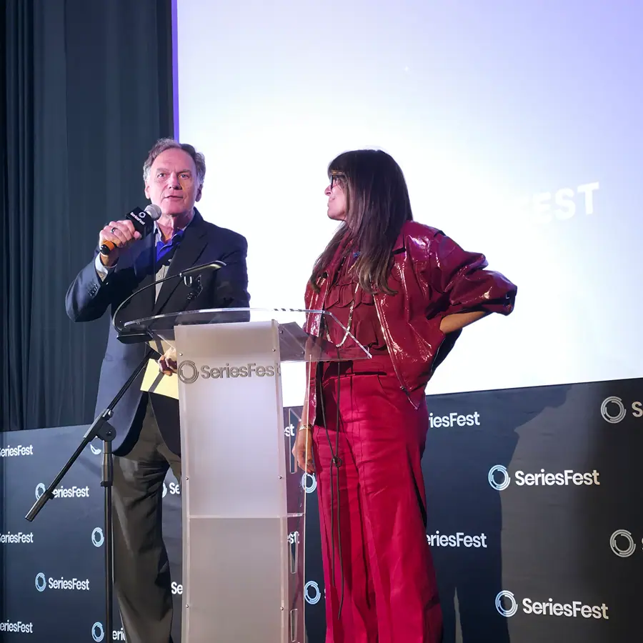 Two presenters at a SeriesFest-branded lectern on the Season 12 stage — one speaking into a handheld microphone, the other standing beside him in a red suit — in front of a step-and-repeat wall of repeated SeriesFest marks, a lit projection screen filling the wall behind them.
