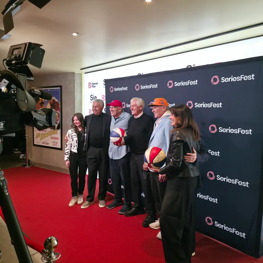 Six people lined up on the red carpet at the SeriesFest step-and-repeat outside the Sie FilmCenter, two of them holding red-white-and-blue ABA basketballs, a broadcast camera in the foreground.