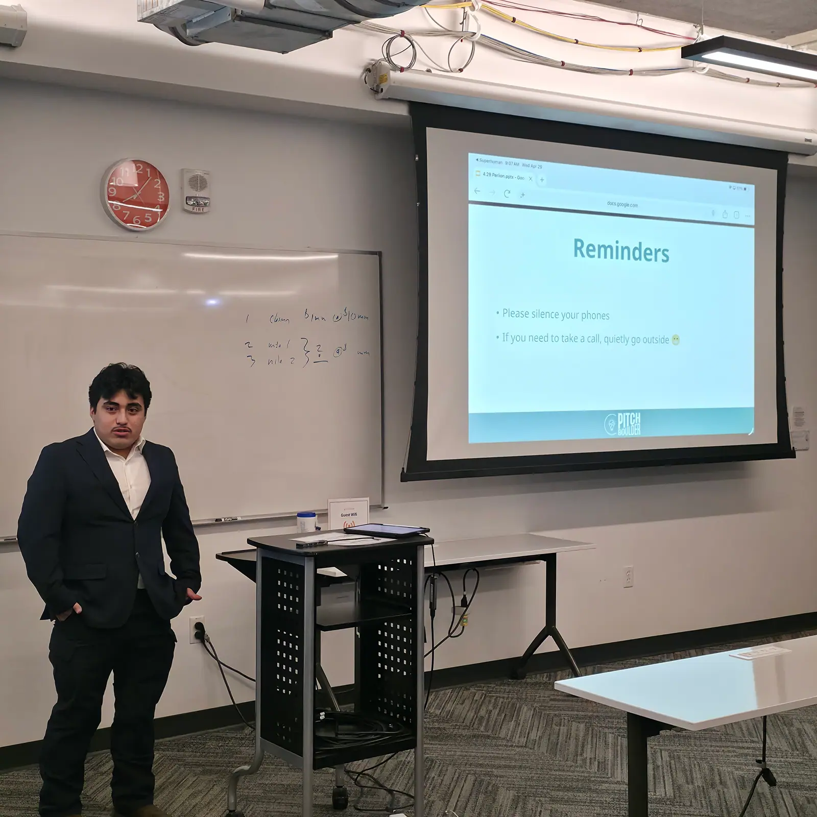 A founder in a blazer taking the floor beside a projected slide at a PitchBoulder session.