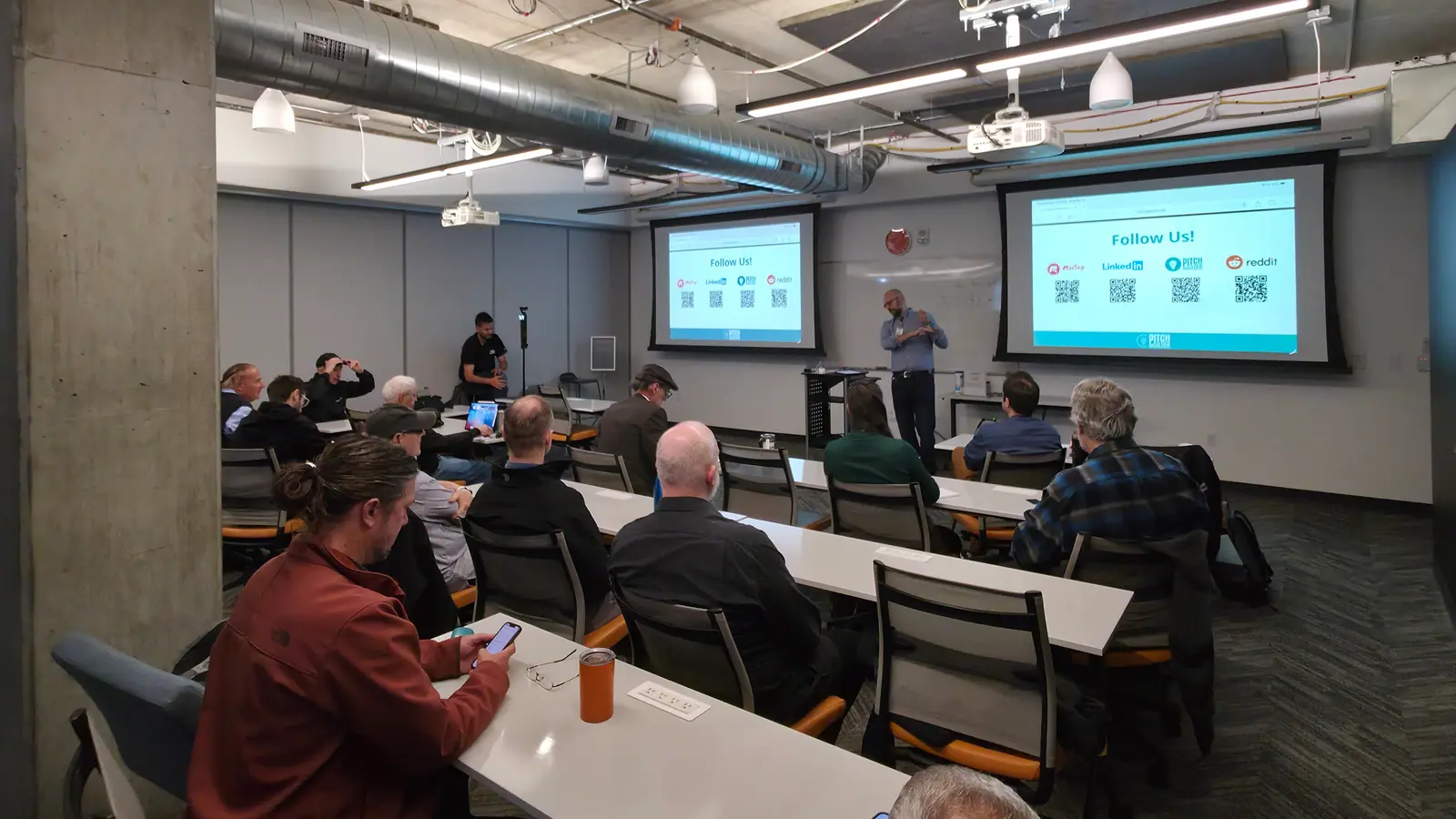 The PitchBoulder room from the back — attendees at long tables, the host on the mic, and the session's “Follow Us!” slide with its QR codes on both projector screens.