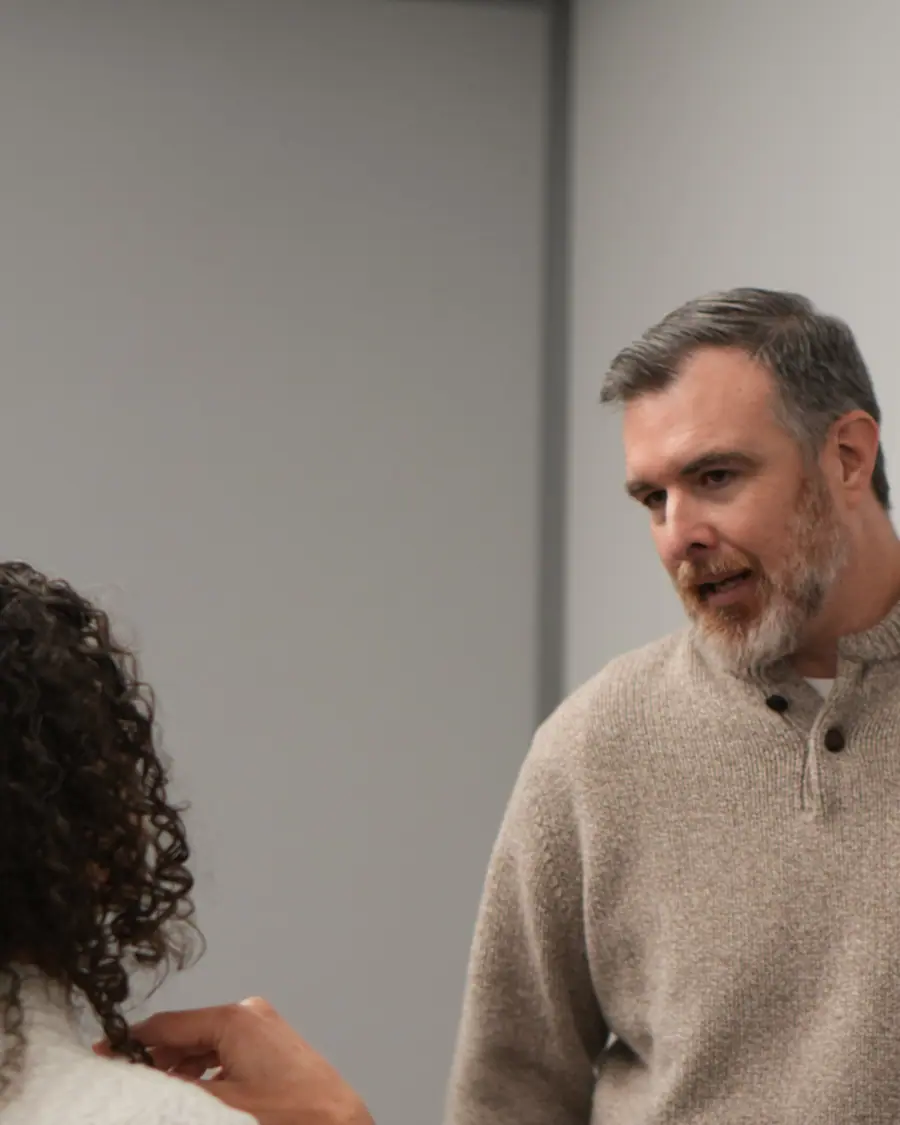 A founder mid-sentence, answering one person one-to-one after his pitch, the listener's hair filling the foreground and a plain grey wall behind.