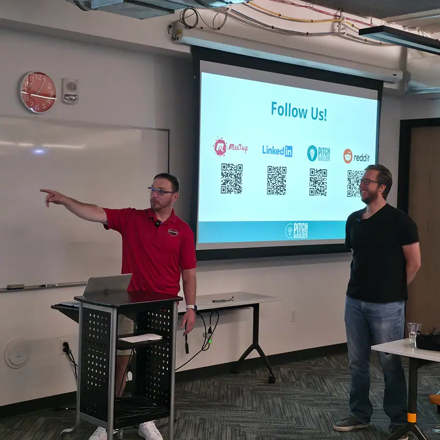 Two men stand together in front of the 'Follow Us!' QR slide, one gesturing toward the room, the other laughing.