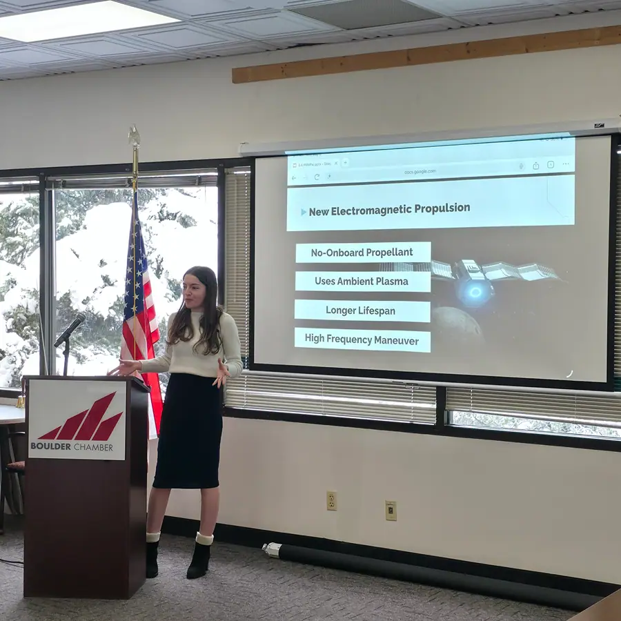 Ashle Jantzen, founder of Leyden Space, mid-gesture at a Boulder Chamber podium, both arms spread wide, a slide reading 'New Electromagnetic Propulsion' projected behind her.