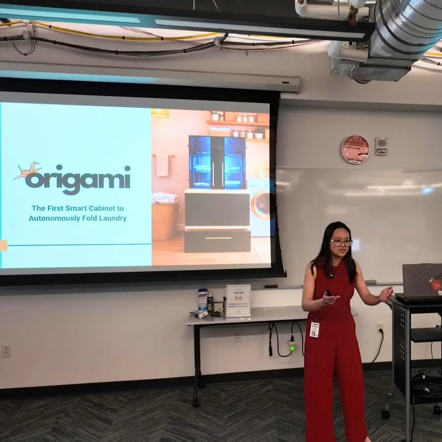 A founder in red presents beside a projected title slide reading 'origami — The First Smart Cabinet to Autonomously Fold Laundry,' with a rendering of the cabinet beside the text.
