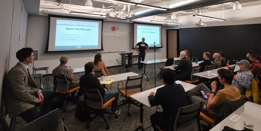 The PitchBoulder room from the back, looking square-on at the front wall — roughly eighteen attendees seated either side of a centre aisle, the host standing between two projector screens that both read “About PitchBoulder”.