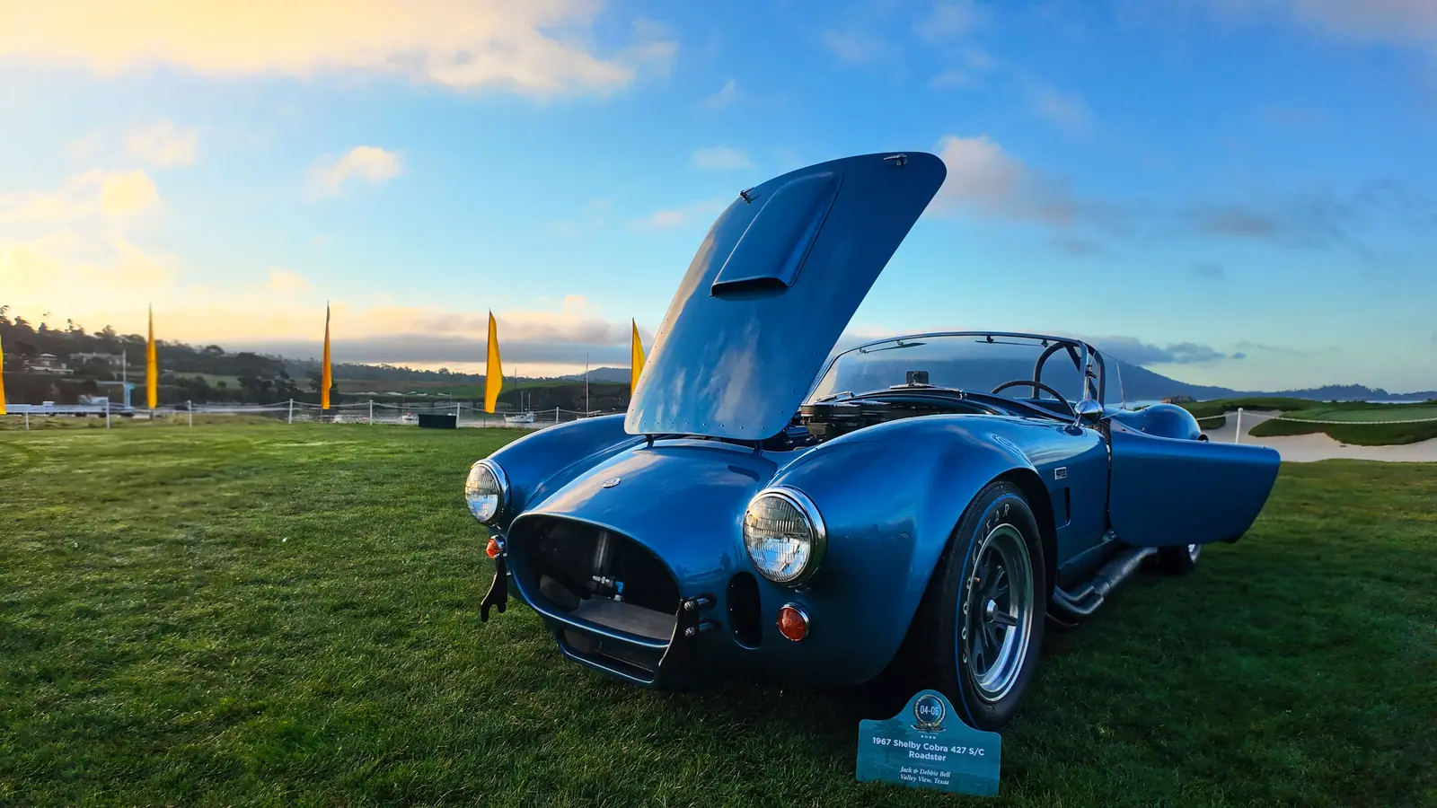 The restored blue 1967 Shelby Cobra 427, hood raised, on the show lawn at sunrise with the coastline behind