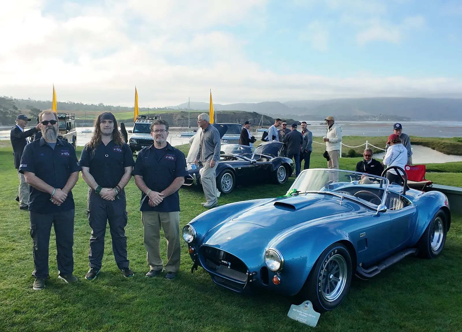 The three-person crew beside the blue Cobra, a silver Cobra and the bay behind
